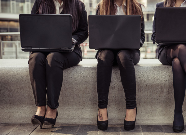 Drei Frauen sitzen mit Laptops auf einer Mauer und arbeiten.