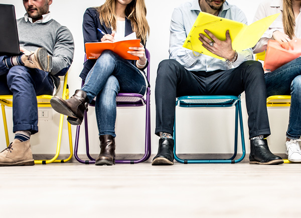 Das Bild zeigt eine Reihe von Personen, die auf bunten Stühlen sitzen und auf etwas warten